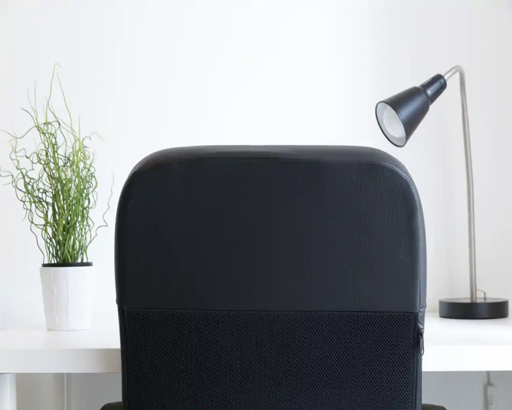 Close-up of a modern black office chair back positioned at a white desk with a small potted plant and a desk lamp, showing a clean, minimal workspace corner.