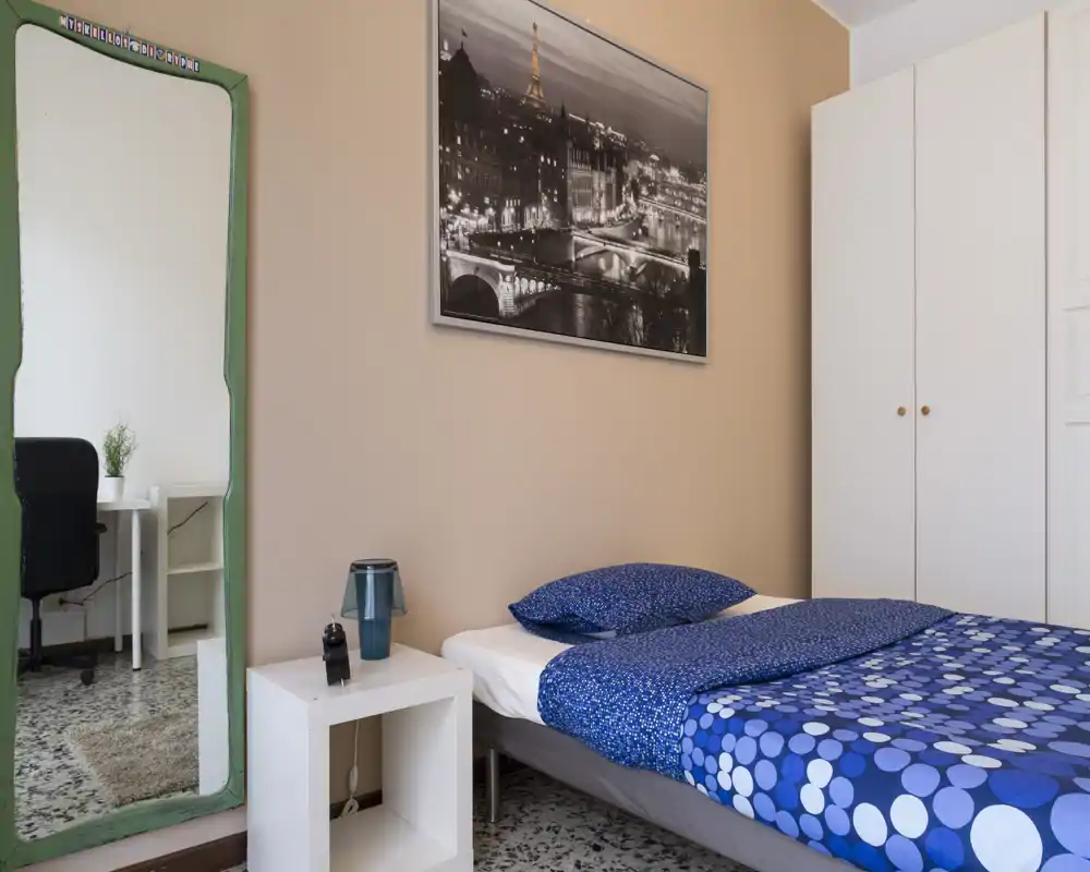 Bright, tidy bedroom with a single bed dressed in blue patterned linens, a bedside table with lamp, a large mirror and a white wardrobe.