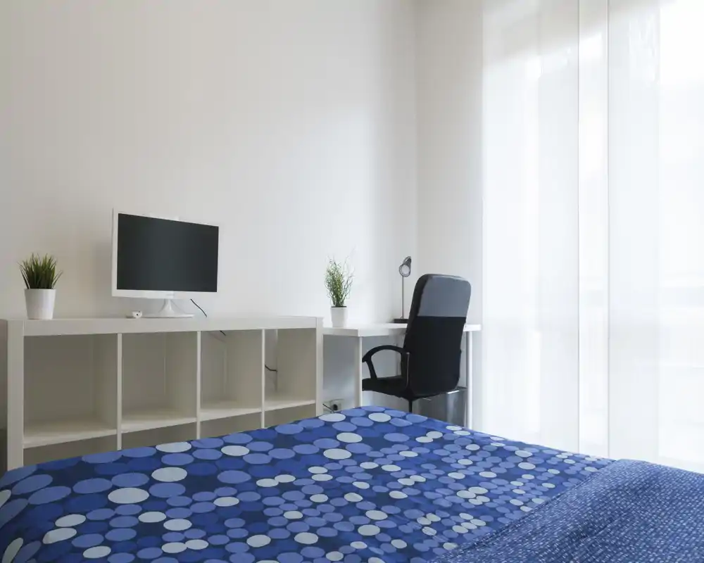 Bright modern bedroom with a patterned blue bedspread, white storage shelving, a desk and chair by a large window letting in abundant natural light.