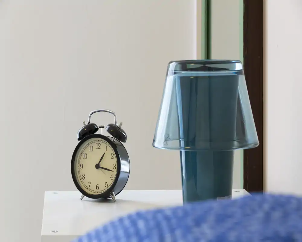 Close-up of a bedside table with a retro alarm clock and a modern translucent lamp; a blue bedcover is slightly visible in the foreground, suggesting a bedroom.