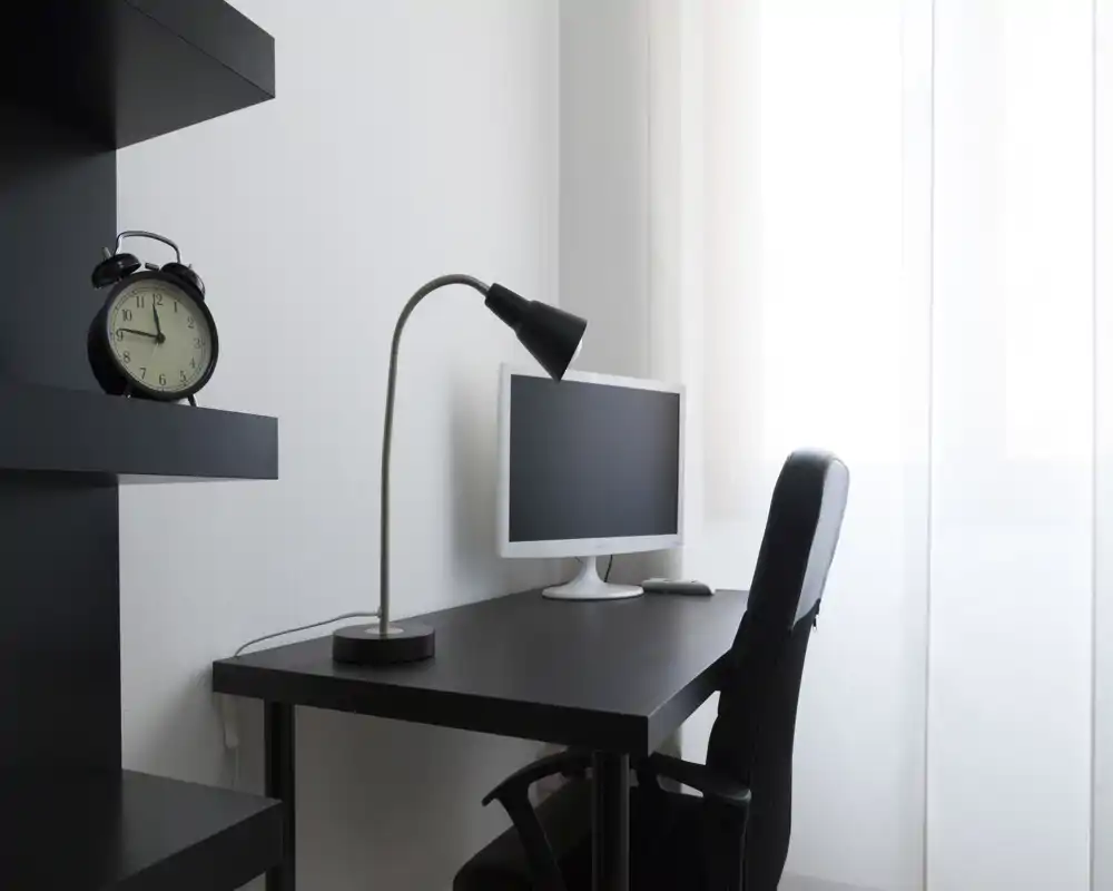 A minimalist workspace corner with a black desk, desk lamp, monitor and office chair beside a bright window.