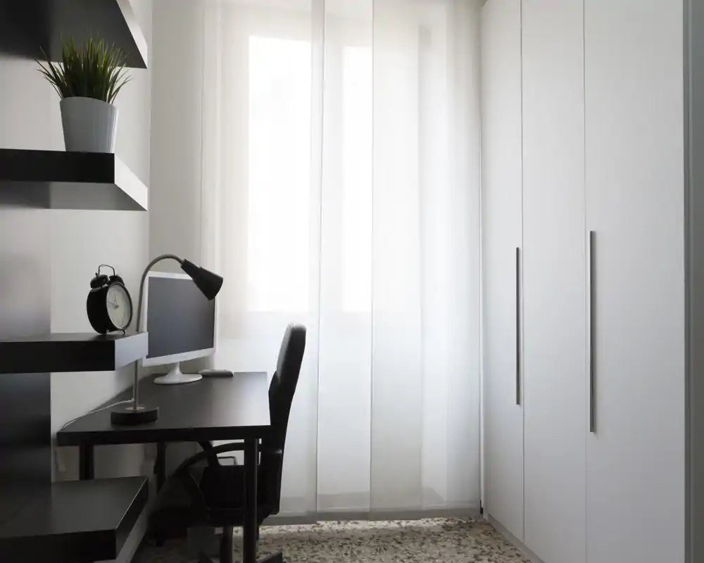 Bright minimalist home office corner inside a living area, featuring a black desk with monitor and lamp, wall shelving and a large white wardrobe beside sheer curtains that diffuse natural light.