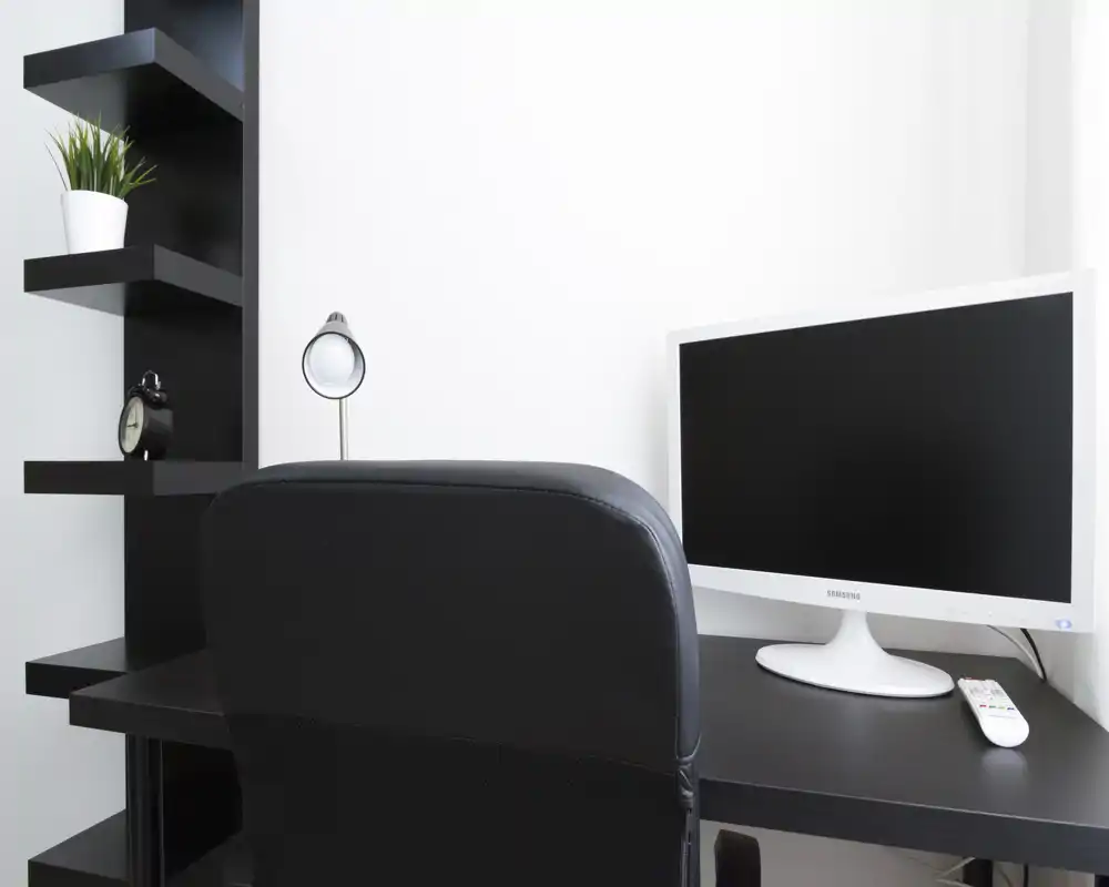 Minimal home workspace with a black desk, ergonomic chair, white monitor and small desk lamp; clean modern styling and a potted plant on shelving.