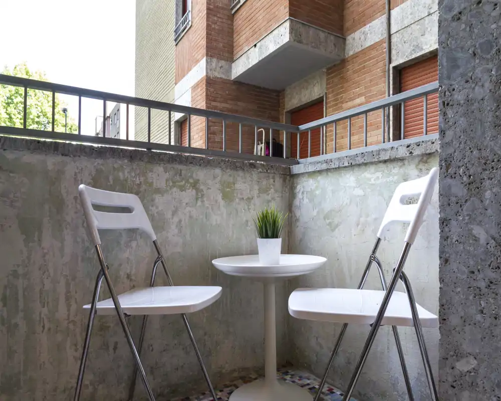 Piccolo balcone urbano con due sedie pieghevoli bianche, un tavolino rotondo e una pianta in vaso — perfetto per un caffè mattutino in uno spazio esterno compatto.