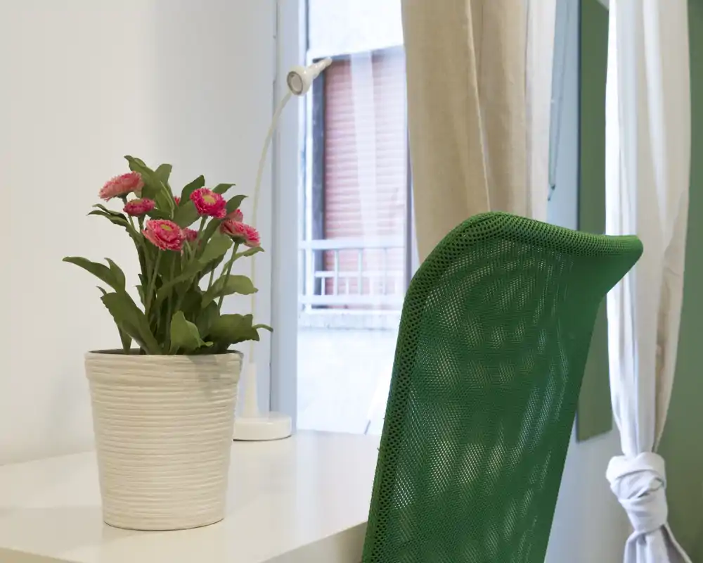 Angolo lavoro luminoso con scrivania bianca, sedia da ufficio verde e vaso con fiori vicino a una finestra con tenda.