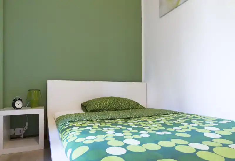 Camera da letto compatta e luminosa con testiera bianca e biancheria a motivo verde. Un piccolo comodino con sveglia e lampada verde coordina lo spazio rendendolo moderno e accogliente.