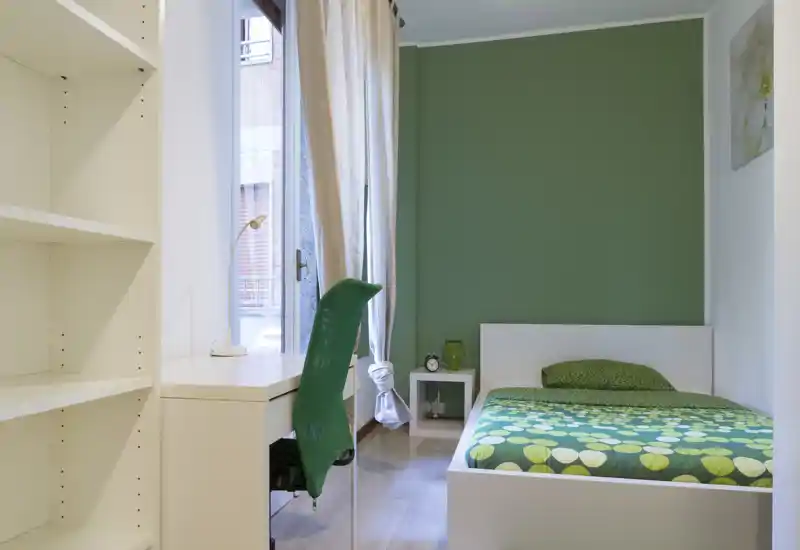 Camera singola luminosa e moderna con parete verde, letto ben rifatto e scrivania compatta vicino alla finestra — ideale per uno studente o un viaggiatore singolo.