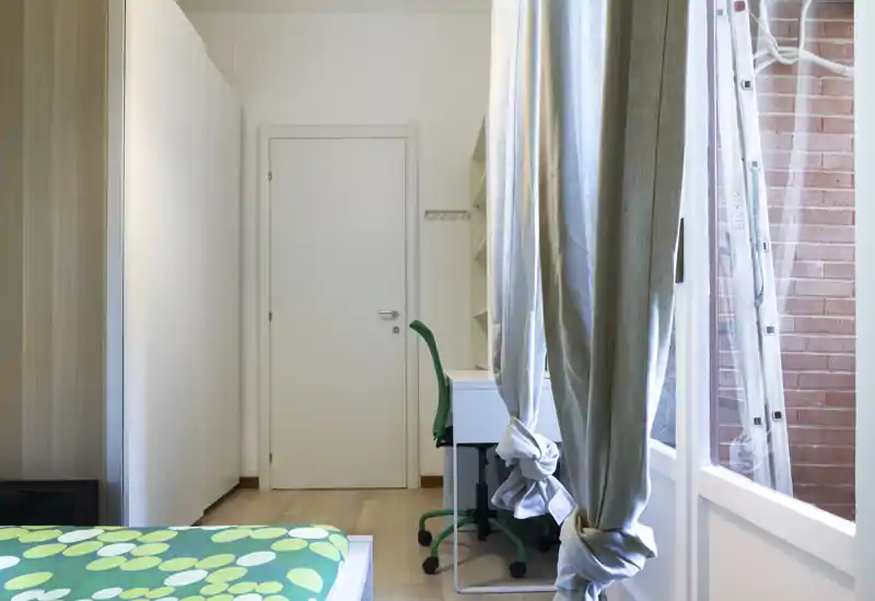 Camera da letto luminosa e ordinata con angolo del letto visibile, copriletto a fantasia e una piccola scrivania vicino alla finestra; luce naturale e finiture neutre per un’atmosfera moderna.