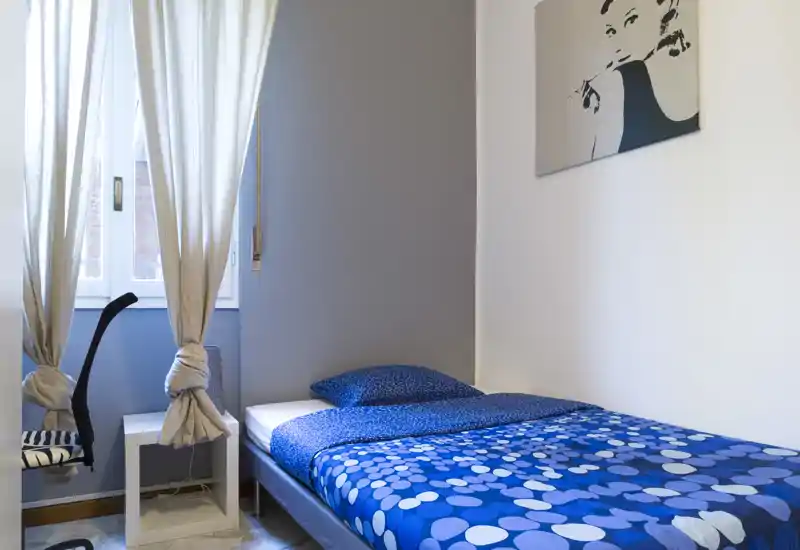 Bright, neatly styled single bedroom with patterned blue bedding, pillow and a small bedside area by a curtained window — ideal for a rental listing emphasizing cleanliness and natural light.