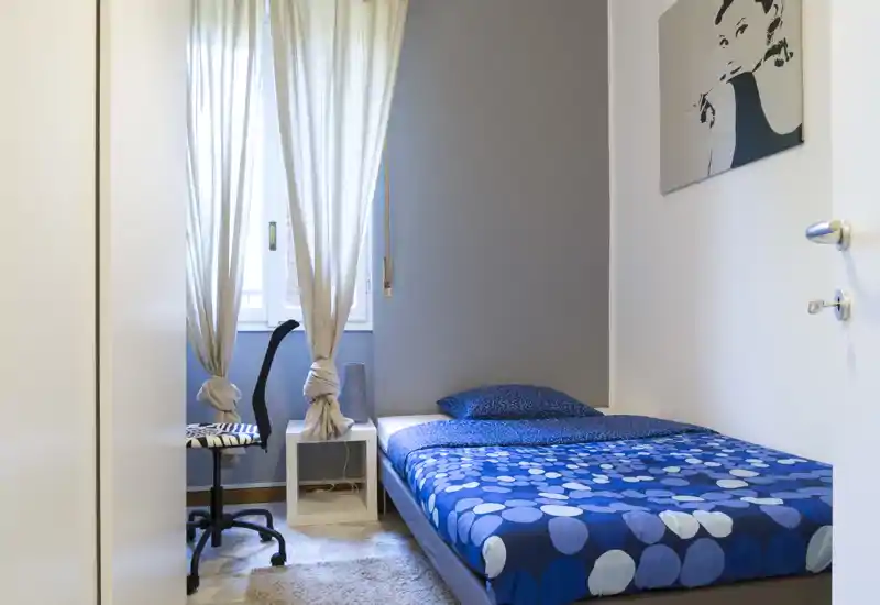 Compact, well-lit bedroom with a double bed dressed in blue patterned bedding, a bedside table and a window with soft curtains providing natural light.