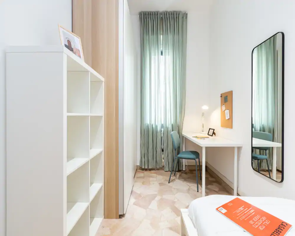 Dormitorio luminoso y minimalista con una cama en primer plano, un escritorio junto a la ventana y cortinas verdes que suavizan la luz.