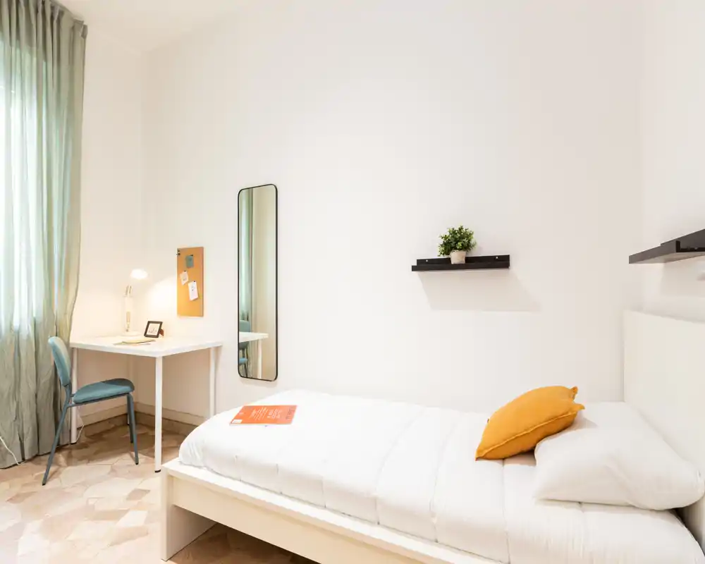 Dormitorio individual luminoso y minimalista con cama blanca, cojín mostaza, escritorio junto a la ventana, espejo de cuerpo entero y estantes sencillos — ideal para estudiante o viajero solo.