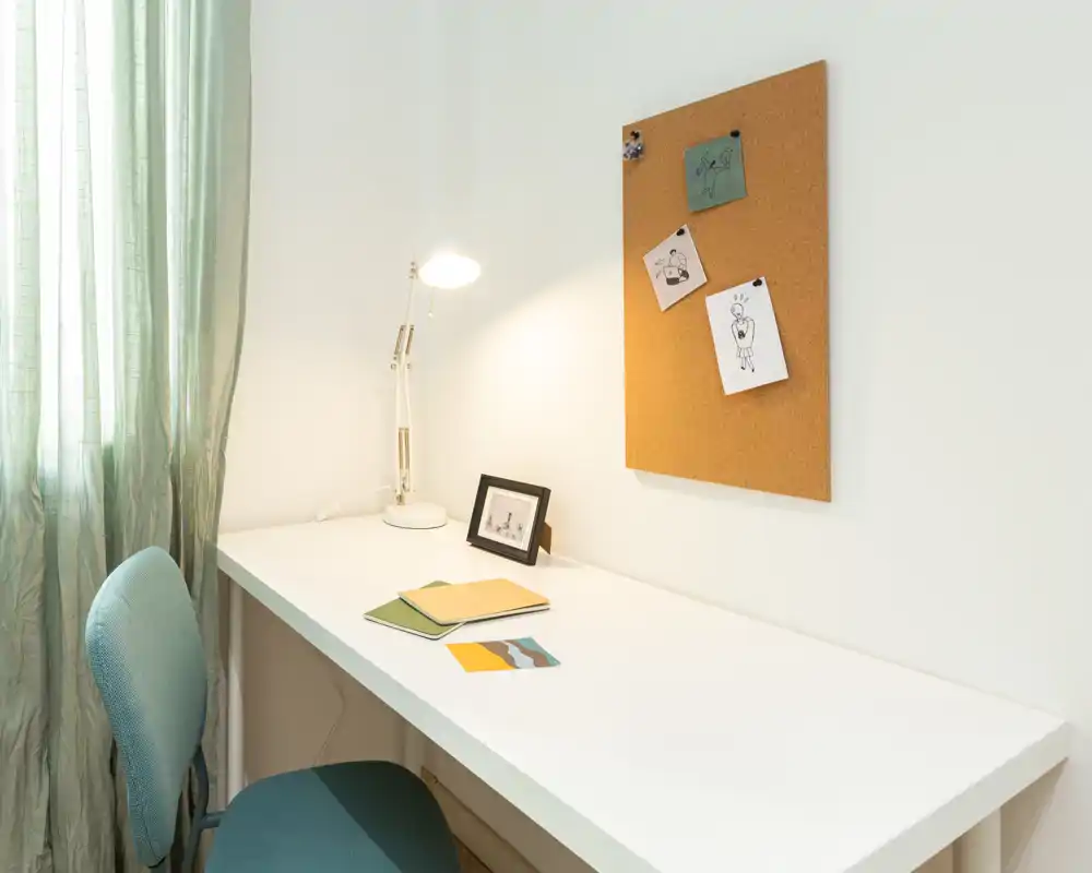 Espacio de trabajo doméstico luminoso y minimalista con escritorio blanco, silla turquesa, lámpara de mesa y corcho — ideal para estudiar o teletrabajar.