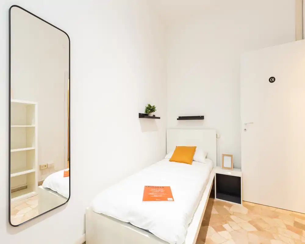 Habitación individual luminosa y minimalista con cama bien hecha, cojín de color cálido y espejo de cuerpo entero que aporta profundidad. Ideal para un huésped individual o alquileres cortos.