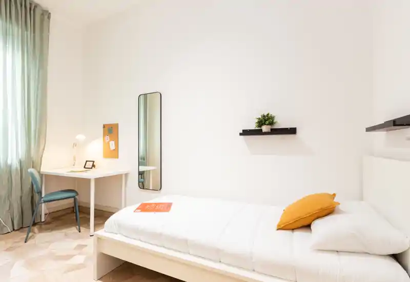 Chambre simple lumineuse et minimaliste avec lit blanc, coussin moutarde, bureau près de la fenêtre, miroir en pied et étagères murales — idéale pour étudiant ou voyageur seul.