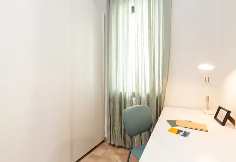 Coin bureau compact et lumineux avec un bureau blanc, une chaise bleu-vert et une lampe près d'une fenêtre aux rideaux légers.
