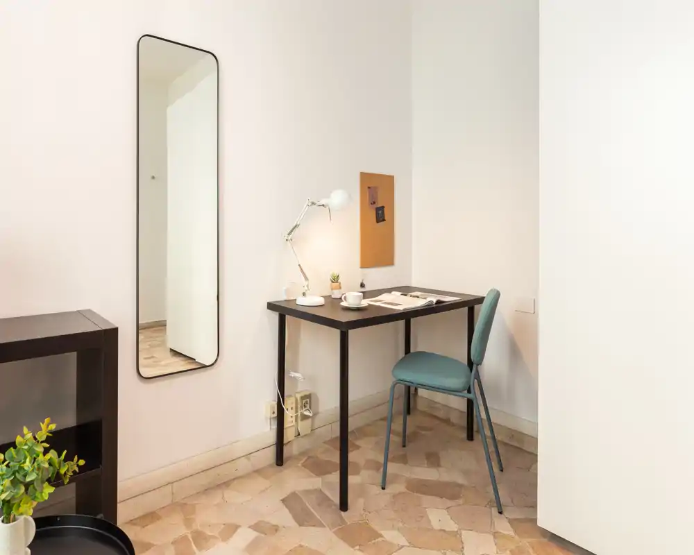 Compact, well-lit living area corner set up as a small workspace: a simple desk with a lamp, chair and wall mirror creates a tidy, functional nook.