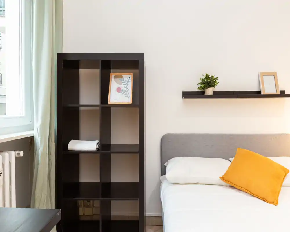 Bright, minimal bedroom with a grey upholstered headboard, white bedding and a warm yellow accent cushion; a black shelving unit and floating shelf with small decor create a tidy, modern look.
