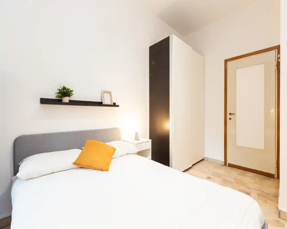 Bright, minimal bedroom with a double bed, neutral headboard and a yellow accent cushion; bedside table and wardrobe visible—ideal for rental listings highlighting a clean, modern sleeping area.