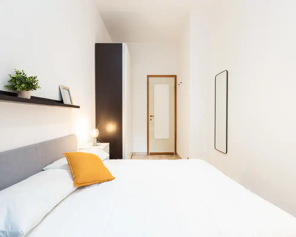 Bright, minimalist bedroom with a large white bed, mustard accent pillow, bedside table and lamp, a tall wardrobe near the door and a wall mirror — clean lines and soft natural light.