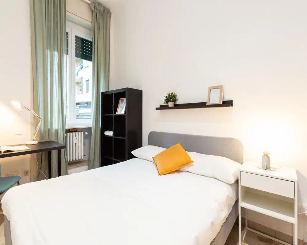 Bright, well-staged bedroom with a double bed, bedside table and desk by the window. Neutral tones, a yellow accent cushion and soft lighting create a welcoming atmosphere ideal for listings.