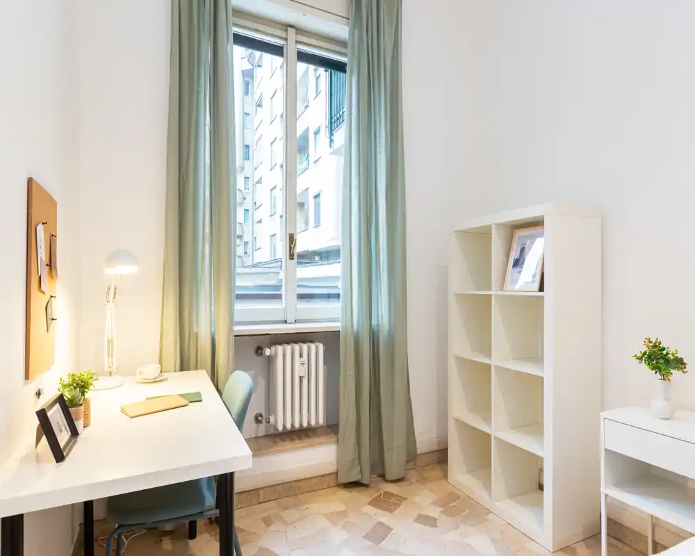 Bright, minimal living/working corner with a desk, chair, lamp and shelving by a large window — ideal for a compact studio or home office.