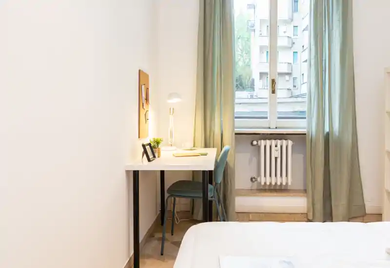 Bright, minimal bedroom corner with a visible bed edge, a white desk with lamp and chair next to a tall window with light green curtains.