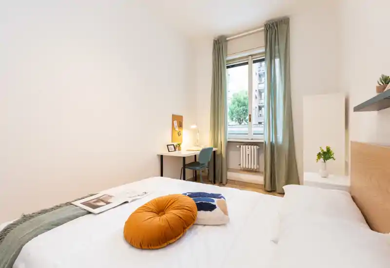 Bright, neatly styled bedroom with a comfortable double bed, decorative cushions and a small desk by the window letting in natural light.