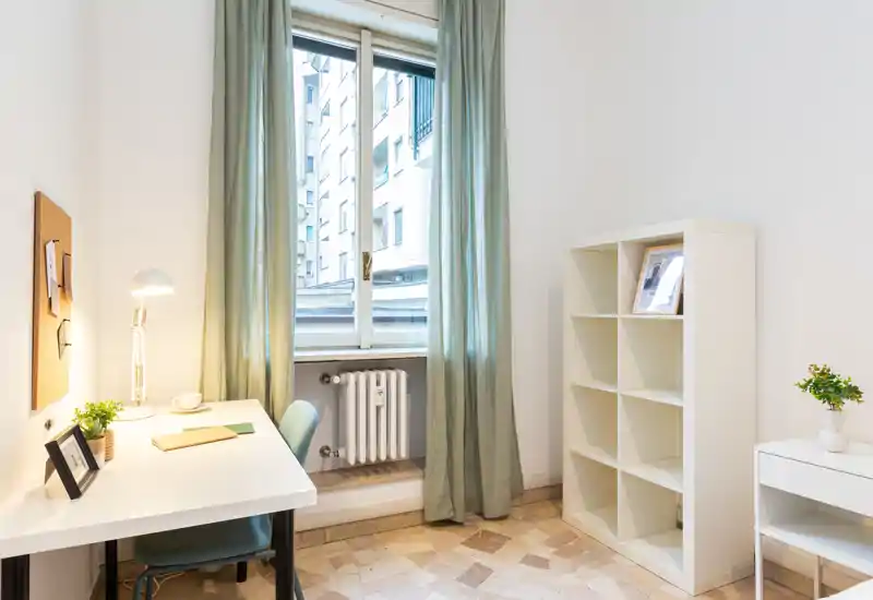 Bright, minimal living/working corner with a desk, chair, lamp and shelving by a large window — ideal for a compact studio or home office.