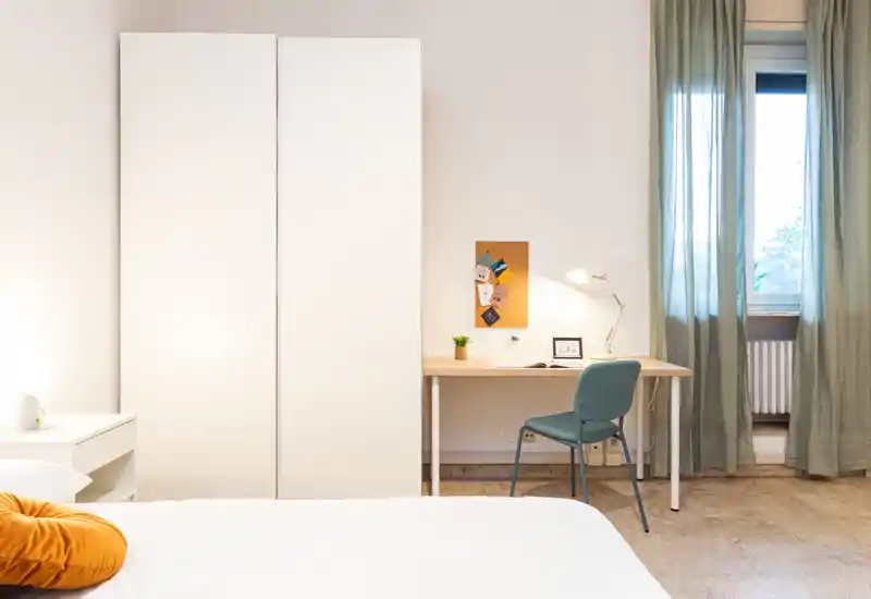 Bright, neatly staged bedroom with a comfortable bed in the foreground, a tall white wardrobe, and a small desk by the window — ideal for restful stays or remote work.