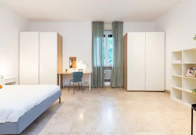 Spacious, modern bedroom with a neatly made bed on the left, two tall wardrobes, a desk by the window and soft natural light creating a calm, airy atmosphere.