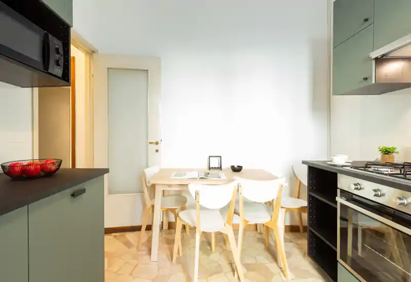 Cuisine moderne et lumineuse avec une petite table en bois et quatre chaises, four encastré et plaque gaz, et meubles verts doux — idéale pour des repas conviviaux dans un espace restreint.