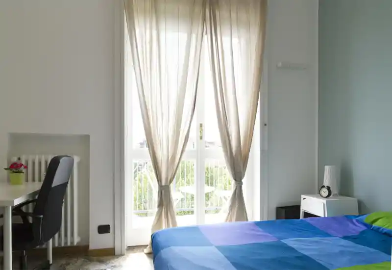Camera da letto luminosa e ordinata con un letto matrimoniale coperto da una coperta blu, un comodino con sveglia e una scrivania vicino alla finestra che lascia entrare molta luce naturale.
