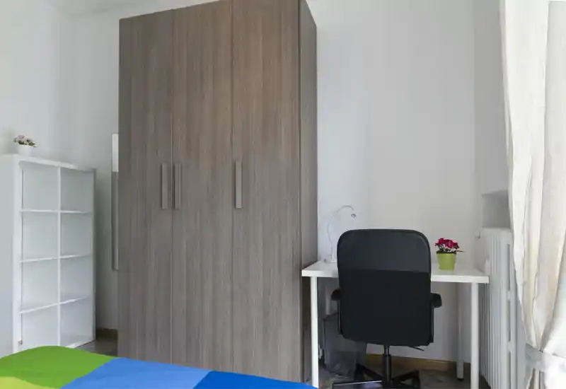 Angolo di camera pulito e semplice con ampio armadio, parte del letto visibile e una piccola scrivania vicino alla finestra — toni neutri e arredo minimale.