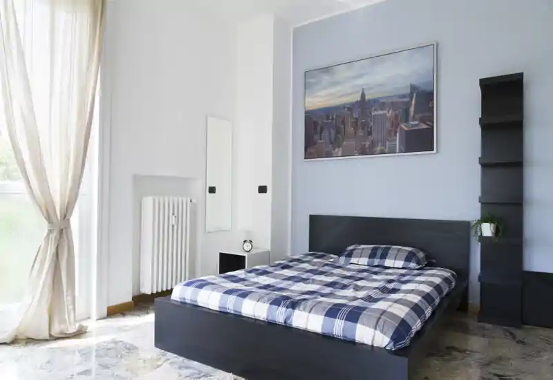 Chambre moderne et lumineuse avec un lit double sur cadre en bois foncé, couette à carreaux, grande fenêtre avec voilages et décoration minimaliste. La lumière naturelle et les lignes épurées offrent un espace accueillant parfait pour une annonce.