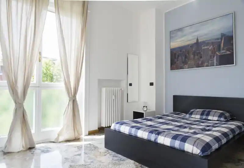 Chambre moderne et lumineuse avec un lit bas noir habillé d'une couette à carreaux bleus, une table de chevet avec réveil et une grande fenêtre aux voilages laissant entrer une belle lumière naturelle.