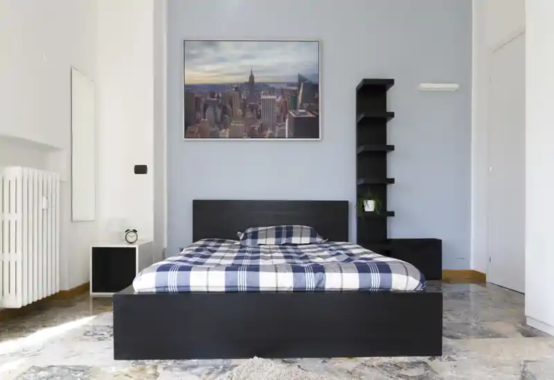 Chambre lumineuse et minimaliste avec un grand lit double en bois foncé, couette à carreaux, table de chevet, étagère et une photo de paysage urbain au-dessus de la tête de lit.