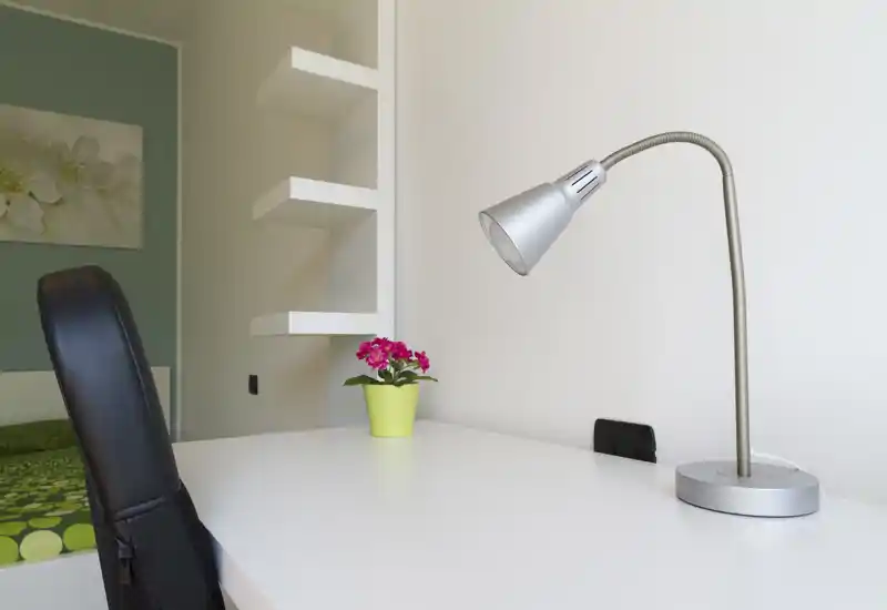 Angolo di camera da letto luminoso e ordinato con una scrivania bianca, lampada moderna e una piccola pianta in vaso; il letto è parzialmente visibile a sinistra, ideale per un ambiente compatto e curato.