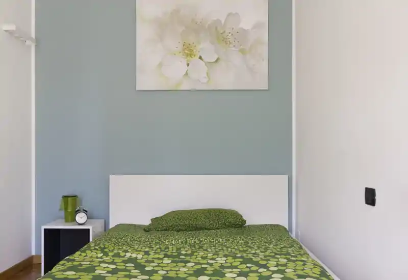 Camera da letto luminosa e semplice, con copriletto verde a fantasia, testiera bianca e quadro sopra; comodino compatto con lampada e orologio.