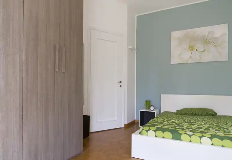 Camera da letto luminosa e dall'arredo semplice con letto piattaforma bianco, biancheria verde a fantasia e comodino; parete color turchese tenue e quadro floreale creano un'atmosfera rilassante.