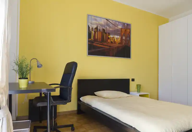 Camera singola luminosa con letto singolo, scrivania e armadio. Pareti gialle calde, quadro con skyline e una pianta creano un ambiente accogliente e luminoso, perfetto per studenti o professionisti single.