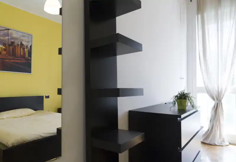 Camera da letto moderna e luminosa con parete gialla, letto basso e luce naturale dalla finestra con tenda; arredi minimal e una pianta rendono l'ambiente fresco.