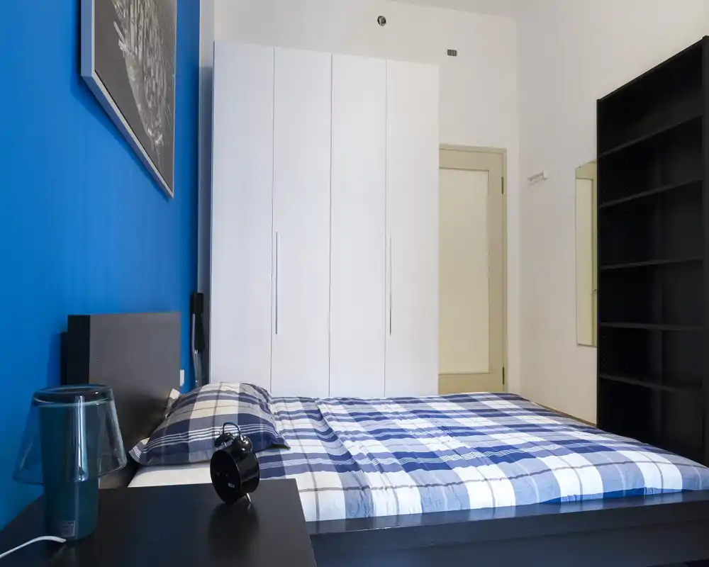 Camera da letto moderna e luminosa con letto matrimoniale con copriletto a quadri blu, armadio bianco a muro e scaffalatura nera. Linee pulite e parete blu accentuata creano un aspetto contemporaneo.