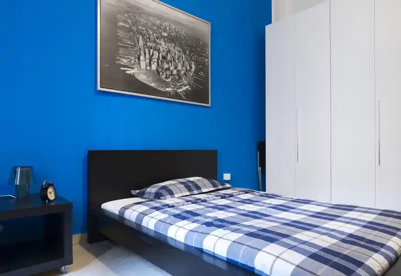 Camera da letto moderna e luminosa con letto matrimoniale con copriletto a quadri blu, testata nera, comodino con lampada e sveglia e un armadio bianco: accogliente e curata.