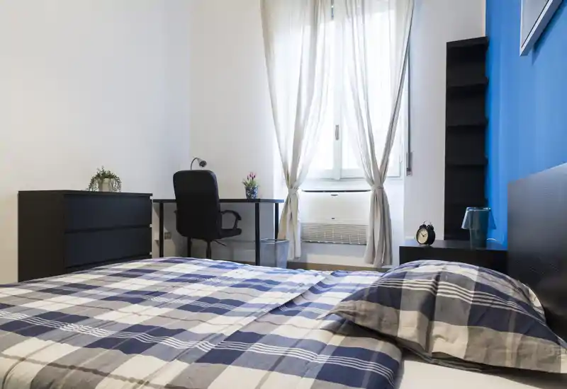 Camera da letto moderna e luminosa con letto matrimoniale con copriletto a quadri blu, scrivania vicino alla finestra e mobili neri minimalisti — perfetta per un soggiorno confortevole.