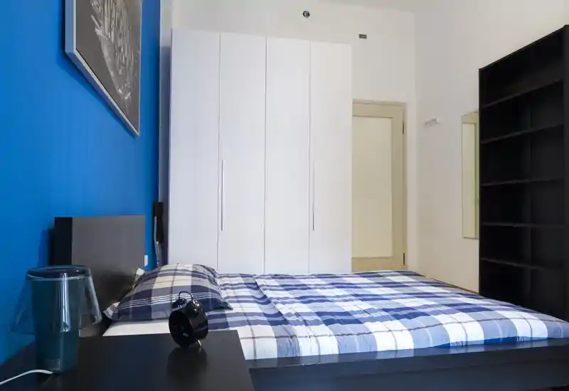 Camera da letto moderna e luminosa con letto matrimoniale con copriletto a quadri blu, armadio bianco a muro e scaffalatura nera. Linee pulite e parete blu accentuata creano un aspetto contemporaneo.