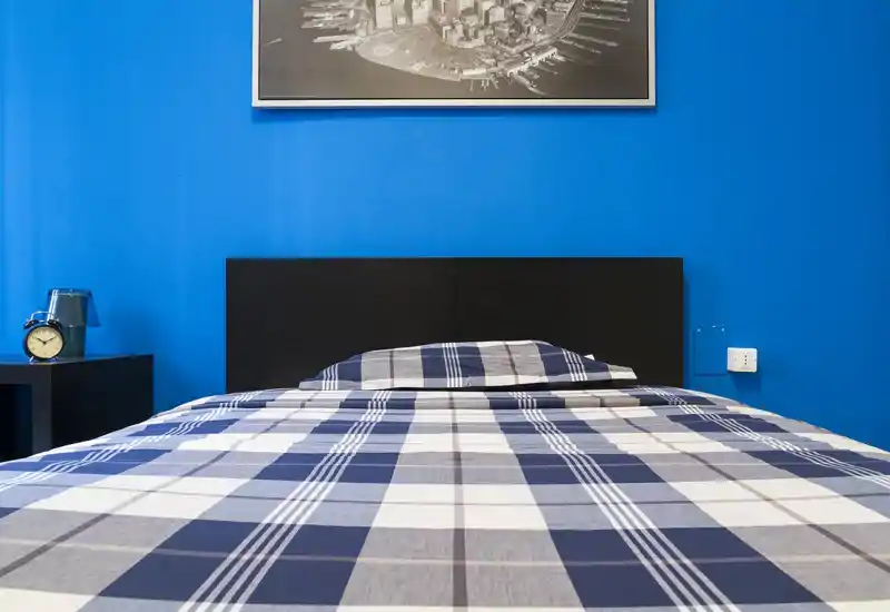 Camera dal piede del letto su una stanza luminosa e ben ordinata; parete blu, biancheria a quadri e comodino conferiscono un aspetto moderno e pulito.