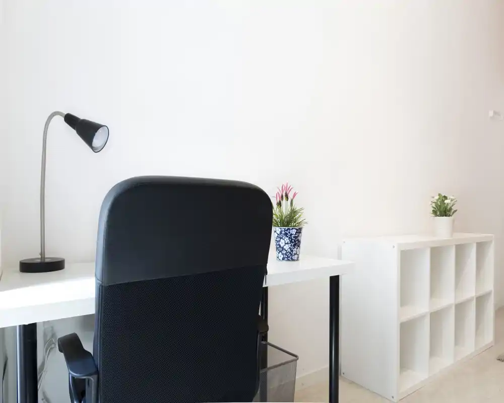 Espacio de trabajo luminoso y minimalista con escritorio blanco, silla ergonómica negra, lámpara de escritorio y estantería abierta. Pequeñas plantas decorativas aportan un toque de vida — ideal para teletrabajo o estudio.