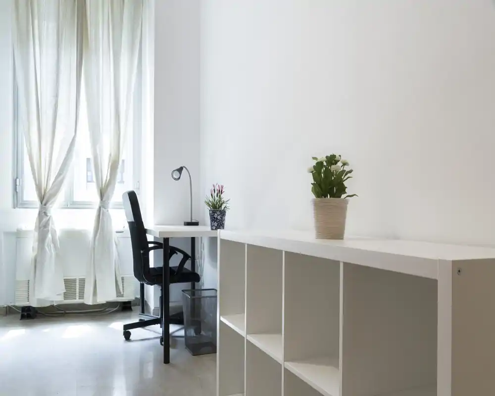 Rincón luminoso y minimalista de salón/oficina con escritorio, silla de oficina y estantería abierta; paredes blancas y luz natural que crean un espacio limpio y funcional.
