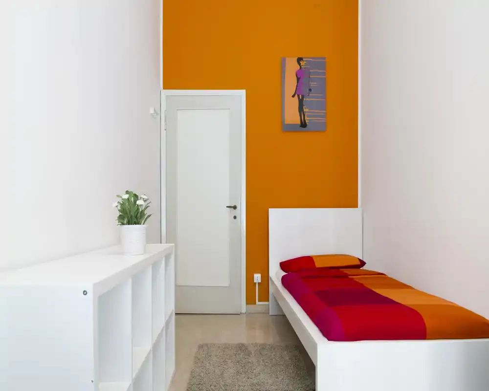 Dormitorio pequeño y luminoso con una cama individual con ropa de cama rojo-naranja, cabecero blanco y estantería blanca. La pared de acento naranja y la decoración minimalista ofrecen un ambiente moderno y acogedor.